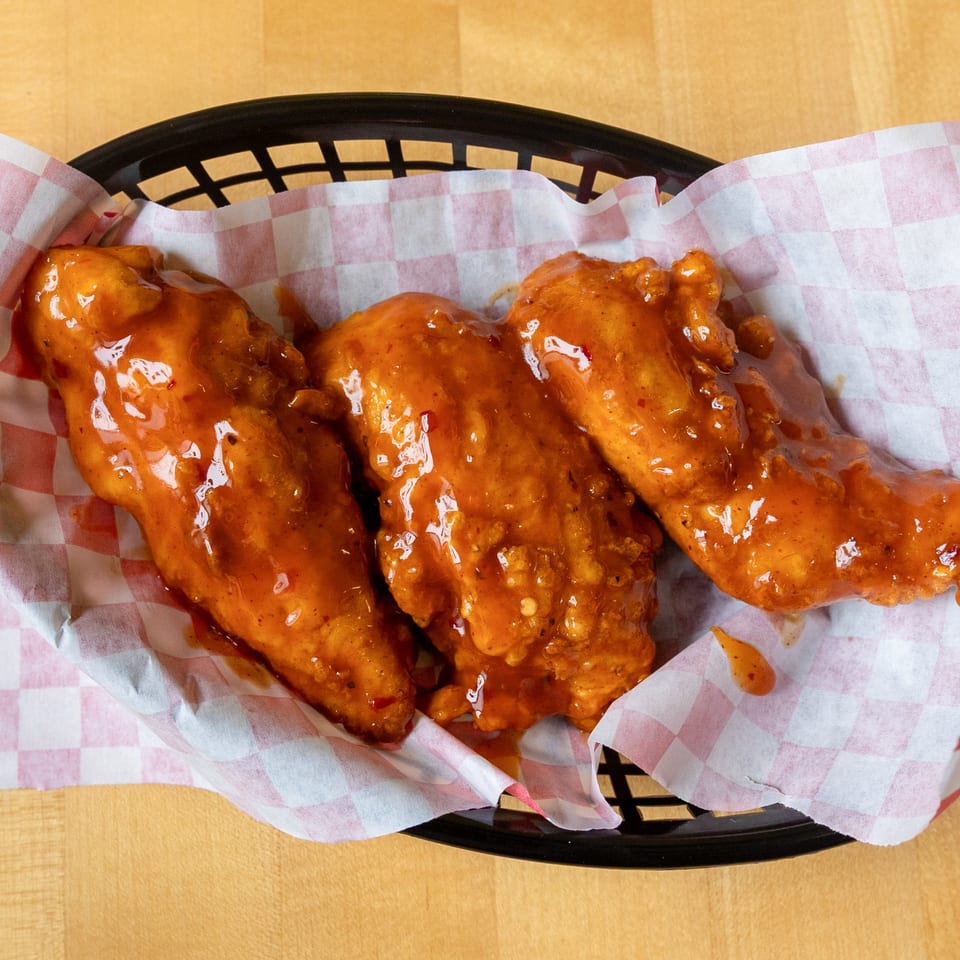 Three WINGSPOT chicken tenders coated in a glossy orange sauce, served in a basket with checkered paper.